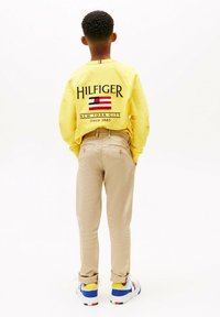 Un sweat-shirt jaune arborant le mot "HILFIGER" et un motif de drapeau, associé à un pantalon beige et des baskets blanches avec des accents bleus et jaunes.