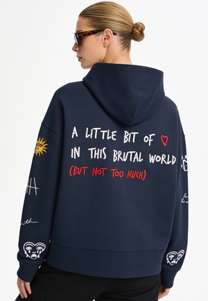 Frau mit Dutt und Sonnenbrille, die einen marineblauen Hoodie mit Text und grafischen Designs trägt, darunter eine Sonne und Herzgesichter auf den Ärmeln.