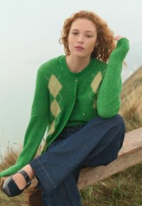 Groene gebreide cardigan met een ruitpatroon in geel, gelaagd over een bijpassend topje. Een paar wijde donkerblauwe jeans en zwarte schoenen.
