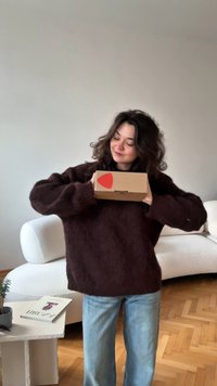 Brązowy, miękki sweter i jasnoniebieskie dżinsy; trzyma małe kartonowe pudełko z czerwonym serduszkiem na naklejce. Jasne, teksturowane tło.