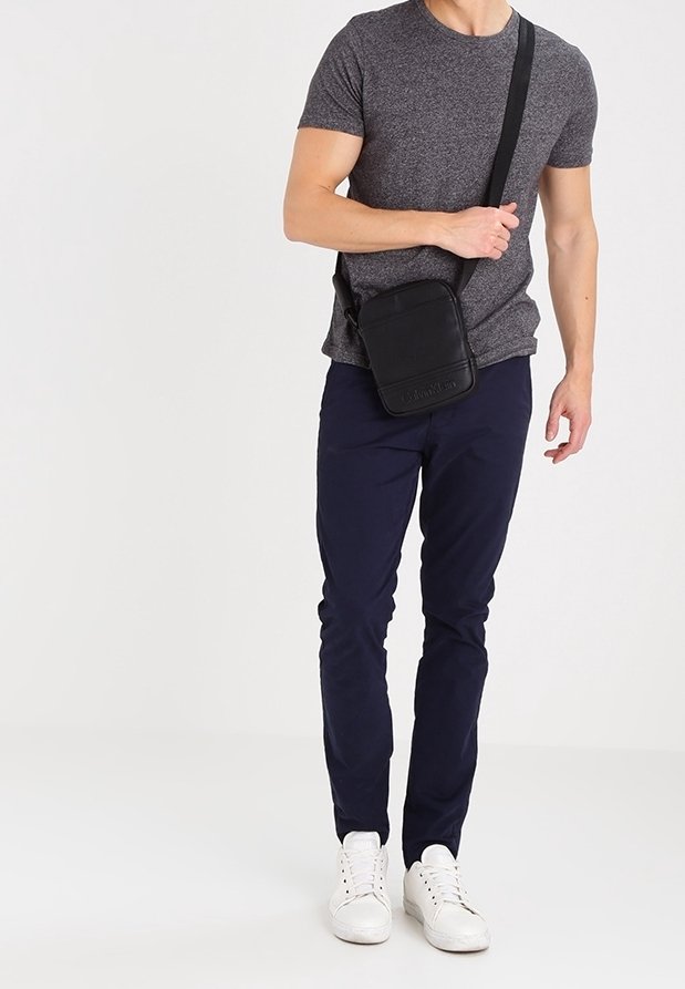 Borsa a tracolla nera in pelle con forma rettangolare semplice, tracolla regolabile e logo impresso nella parte anteriore, indossata con una maglietta grigia e pantaloni blu navy.