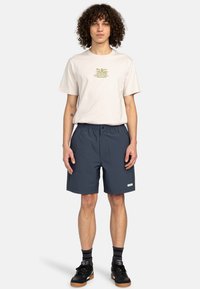 Ljusbeige bomulls-t-shirt med grafiskt tryck, i kombination med marinblå shorts med elastisk midja och sidofickor. Svarta sneakers.