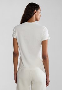 Kurze weiße T-Shirt mit rundem Ausschnitt, glatter Textur und subtilen Rückenetikett-Details, kombiniert mit passenden hellen Hosen.