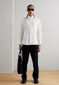 Chemise blanche en lin à manches longues, pantalon noir à jambes larges, lunettes de soleil noires et sac fourre-tout en cuir noir. Textures lisses et design minimaliste.
