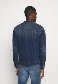 Temno modra jeans jakna z izbledelim vzorcem, z gumbnimi manšetami in blagovno znamko na hrbtni strani. Jakna ima klasičen kroj in ravne šive.