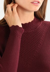Bordeauxroter Strickpullover mit strukturierten vertikalen und Diamantmustern, gerippten langen Ärmeln und einem gewellten Ausschnittdetail.