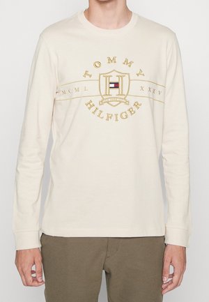 Kremowa koszulka z długim rękawem z złotą haftowaną logo z napisem "Tommy Hilfiger" i wzorem tarczy. Miękki materiał bawełniany.