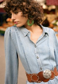 Cămașă din denim cu nasturi în față, mâneci lungi bufante și o curea din piele maro deschis cu elemente circulare argintii și ținte. Cercei verzi rotunzi.