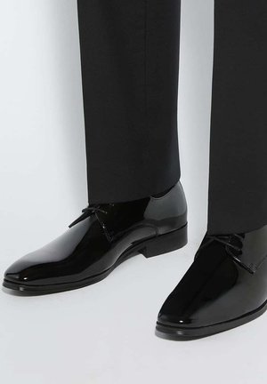 Chaussures de ville noires brillantes portées avec un pantalon de costume noir, présentées sur un fond gris clair.