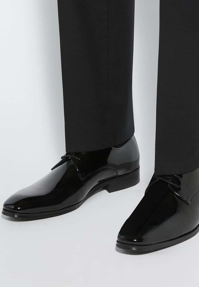Chaussures de ville noires brillantes portées avec un pantalon de costume noir, présentées sur un fond gris clair.