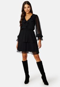 Schwarzes Chiffonkleid mit gerafftem Ausschnitt und langen, durchsichtigen Ärmeln, tailliert, mit schichtigem Rock, kombiniert mit kniehohen schwarzen Stiefeln.