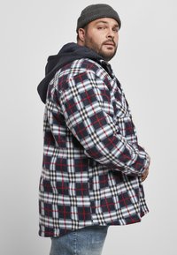 Camisa de franela de cuadros en azul marino, rojo y blanco, con una sudadera con capucha de un azul oscuro en capas. Presenta un cierre de botones en el frente y bolsillos laterales. Textura suave.