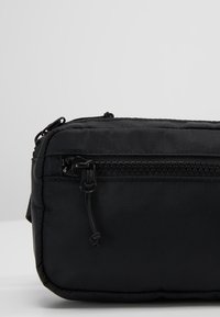 Sac en nylon noir de forme rectangulaire, doté d'une fermeture éclair supérieure et d'une surface en tissu texturé. Les détails incluent une languette de tirette et des coutures.