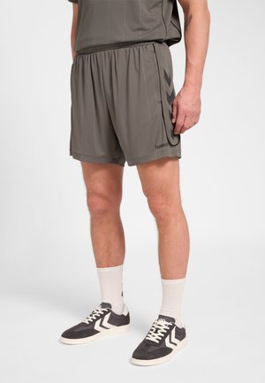 Ein Mann trägt graue Sportshorts, weiße Socken und schwarz-weiße Sneakers und steht vor einem einfarbigen Hintergrund.