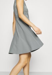 Harmaa mekko, jossa on pitsinen yläosa ja levenevä helma; hihaton malli ja sileä pinta, korostaen kerroksellista kangasta ja lennokasta liikettä.