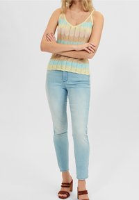 Sweter bez rękawów z falistym wzorem w pastelowych kolorach (niebieski, beżowy, żółty), w połączeniu z jasnymi dżinsami typu skinny i brązowymi sandałami na słupku.