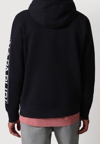 Felpa navy in misto cotone, con cappuccio regolabile, polsini a costine e stampa del logo bianco lungo la manica sinistra. Pantaloni della tuta grigi sotto.