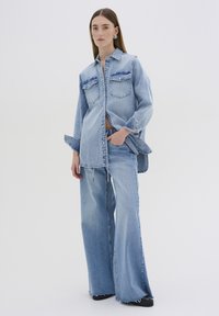 Lys blå denimoutfit med en lang skjorte og brede bukser med slidte kanter, lomme detaljer og synlige sømme.