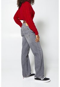 Graue Jeans mit weitem Bein und hoher Taille, Gesäßtaschen und einem roten, gestrickten Cropped-Pullover. Modell trägt schwarze Sneaker. Glatter, lässiger Stil.