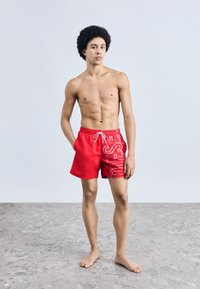 Punased ujumispüksid paelaga vöökohal, küljel julge valge kirjaga. Pehme, sile kangas ja reieluud ületav lõige.