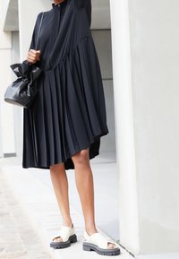 Černé plisované šaty s vysokým výstřihem a krátkými rukávy, v kombinaci s lehkými otevřenými sandály a černou kabelkou.
