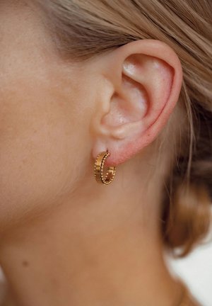 Goldene Creolen mit einer strukturierten Oberfläche und kleinen Perlenakzenten, die am Ohrläppchen hängen, im Hintergrund weiches, hellfarbiges Haar.