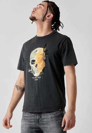T-shirt en coton noir avec un imprimé crâne graphique en jaune et blanc. Il présente un col rond et des manches courtes, associé à un jean gris.