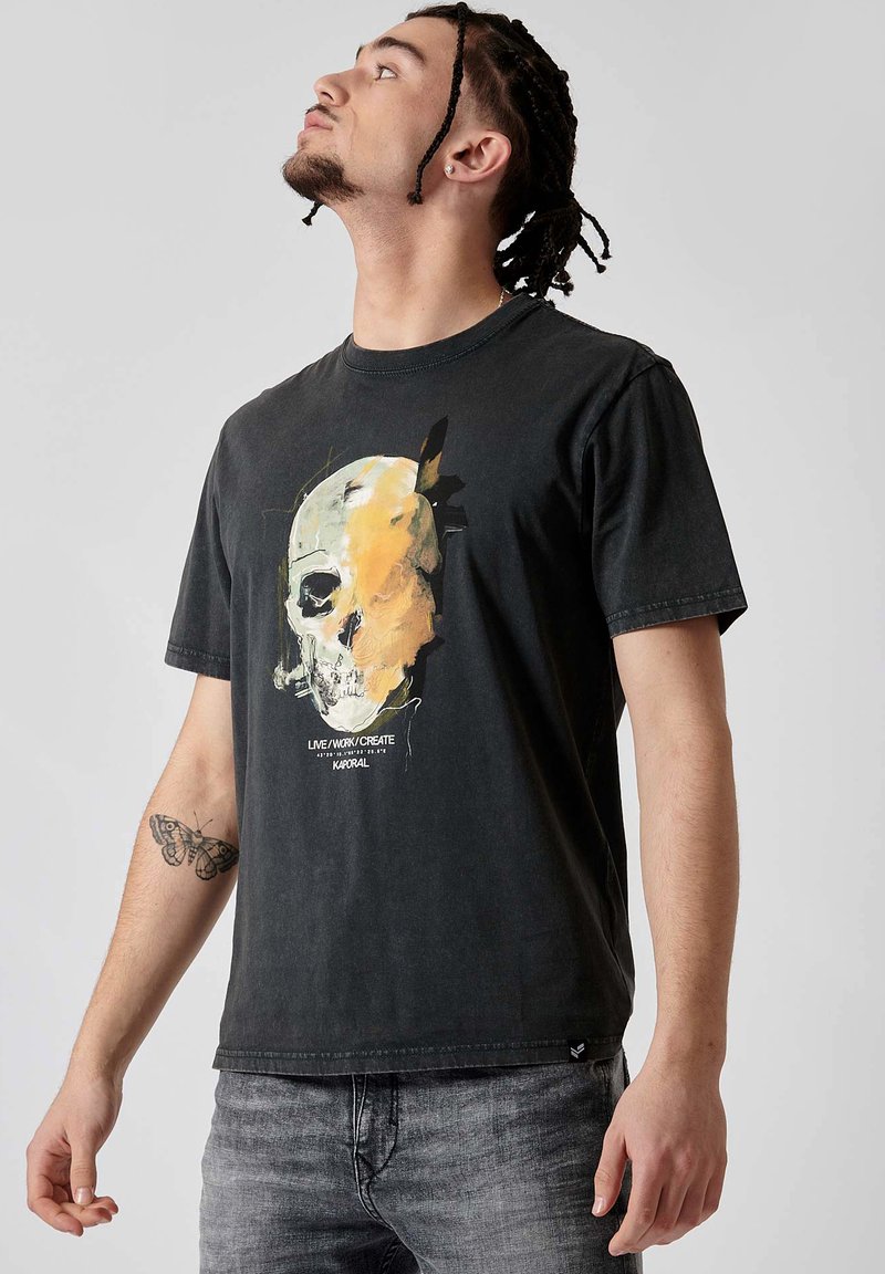 T-shirt en coton noir avec un imprimé crâne graphique en jaune et blanc. Il présente un col rond et des manches courtes, associé à un jean gris.