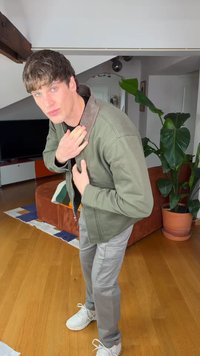 Ung mann justerer kragen på grønn jakke, står i stuen med tregulv, brun sofa, potteplante og TV i bakgrunnen.