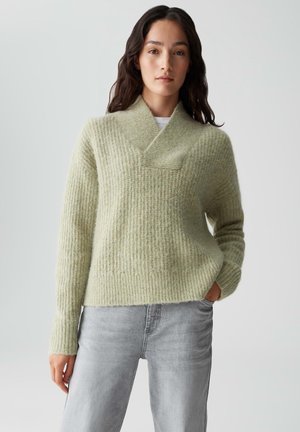 Lichtgroene gebreide trui met een gestructureerd, ribbel patroon, een opgerolde kraag en een relaxed fit, gecombineerd met lichtgrijze denim jeans.