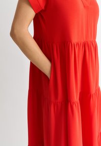 Red tiered dress made of lightweight fabric, featuring short sleeves, a v-neck, and side pockets with a soft, smooth texture.