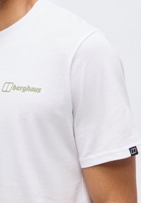 Κοντινό πλάνο ενός ατόμου που φοράει λευκό μπλουζάκι Berghaus με το λογότυπο στο στήθος και μια μικρή ετικέτα στο μανίκι, σε ουδέτερο φόντο.