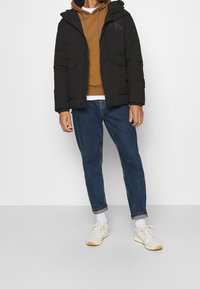 Doudoune noire à capuche, avec poches avant et un logo discret sur la poitrine, portée sur un sweat à capuche marron et un jean bleu, assortie à des baskets blanches.