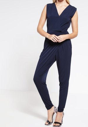 Vrouw draagt een marineblauwe mouwloze jumpsuit in overslagstijl met taps toelopende pijpen en zwarte sandalen met hoge hak en enkelbandjes.