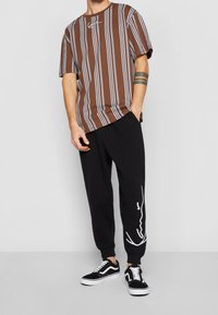 Muž oblečený v tričku s hnědo-bílými vertikálními pruhy, černých joggerách s bílým nápisem na nohavici a černých teniskách s bílými tkaničkami.