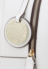 White textured leather bag with a gold circular tag embossed with "Emporio Armani." Features a brown zipper with a gold pull tab.