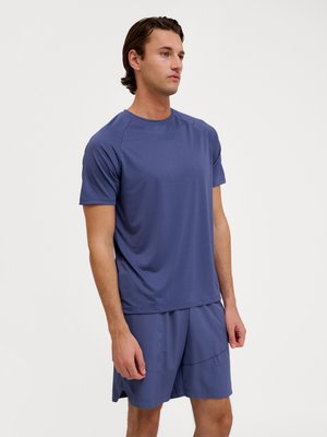 Man staat, draagt een bijpassend blauw sportief T-shirt en korte broek, kijkt lichtjes opzij tegen een effen witte achtergrond.
