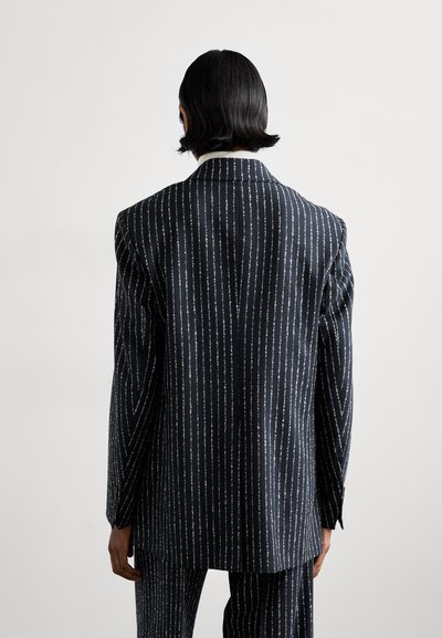 Προσαρμοσμένο μαύρο σακάκι κοστούμι, με ρίγες σε λευκό χρώμα, περιλαμβάνοντας φαρδιές πέτες και δομημένους ώμους. Θέα από πίσω.