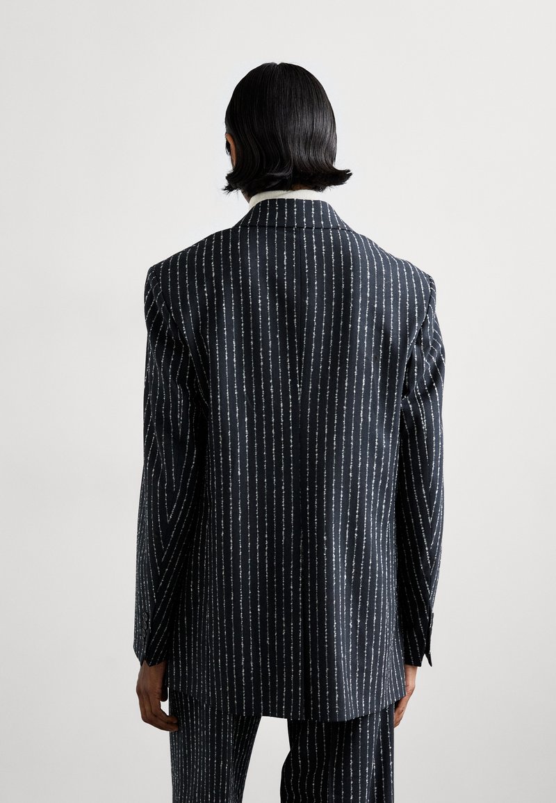 Dopasowana czarna marynarka, w paski z białymi akcentami, z szerokimi klapami i wyraźnymi ramionami. Widok z tyłu.
