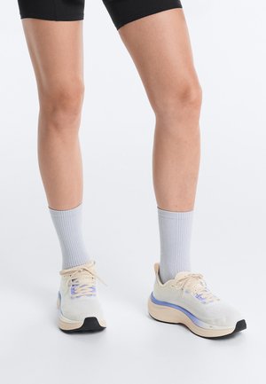 Nogi w czarnych szortach, szarych skarpetach i białych butach do biegania z jasnoniebieskimi i beżowymi akcentami na białym tle.