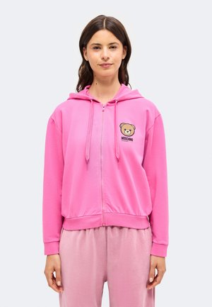 Femme portant un sweat à capuche zippé rose vif avec un logo d’ours en peluche et un pantalon rose clair assorti, debout devant un fond clair uni.