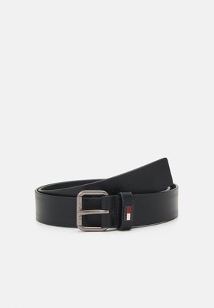 Cinturón de cuero negro con una hebilla de metal plateado. Presenta una pequeña franja de logo en rojo, blanco y azul en la hebilla. Textura suave, diseño clásico.