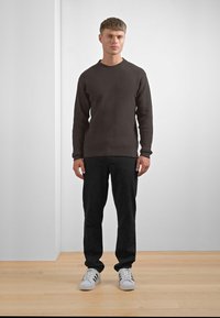Brown ribbed sweater, black pants, white sneakers with black stripes. Model standing in front of a plain backdrop on wooden floor.