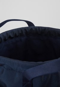 Bolsa de tela azul marino abierta con correa ajustable y borde superior elástico fruncido sobre un fondo gris claro.