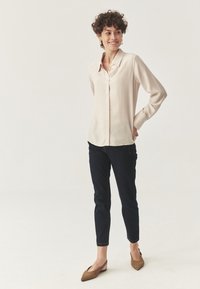 Camicia beige a maniche lunghe con bottoni, dalla texture liscia, abbinata a jeans aderenti scuri e ballerine marroni con punta. 