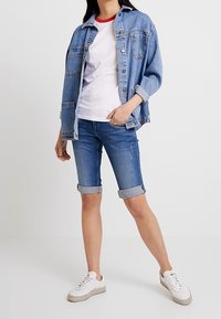 Giubbotto di jeans con lavaggio azzurro chiaro, t-shirt bianca con dettagli rossi e pantaloni shorts con orlo arrotolato. Sneakers bianche completano l'outfit.