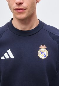 Άντρας που φοράει μπλε σκούρο φούτερ Real Madrid με λευκό λογότυπο Adidas και σήμα συλλόγου στο στήθος, δείχνοντας το κάτω μέρος του προσώπου και τον λαιμό.