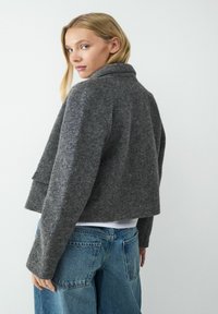 Szara wełniana kurtka z kołnierzykiem, krótki fason i długie rękawy. Materiał ma teksturowane wykończenie, w połączeniu z jasnoniebieskimi szerokimi dżinsami.