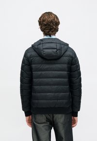 Musta untuvatakki hupulla, jossa on tikattu kuvio ja teksturoitu pinta. Takki on alareunastaan kontrastimateriaalia oleva, tumma kangaspaneeli.