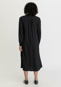 Schwarzes Langarmkleid mit geradem Schnitt, ausgestattet mit einer gerafften Rückenpartie und knopfbaren Manschetten, aus glattem Stoff gefertigt.